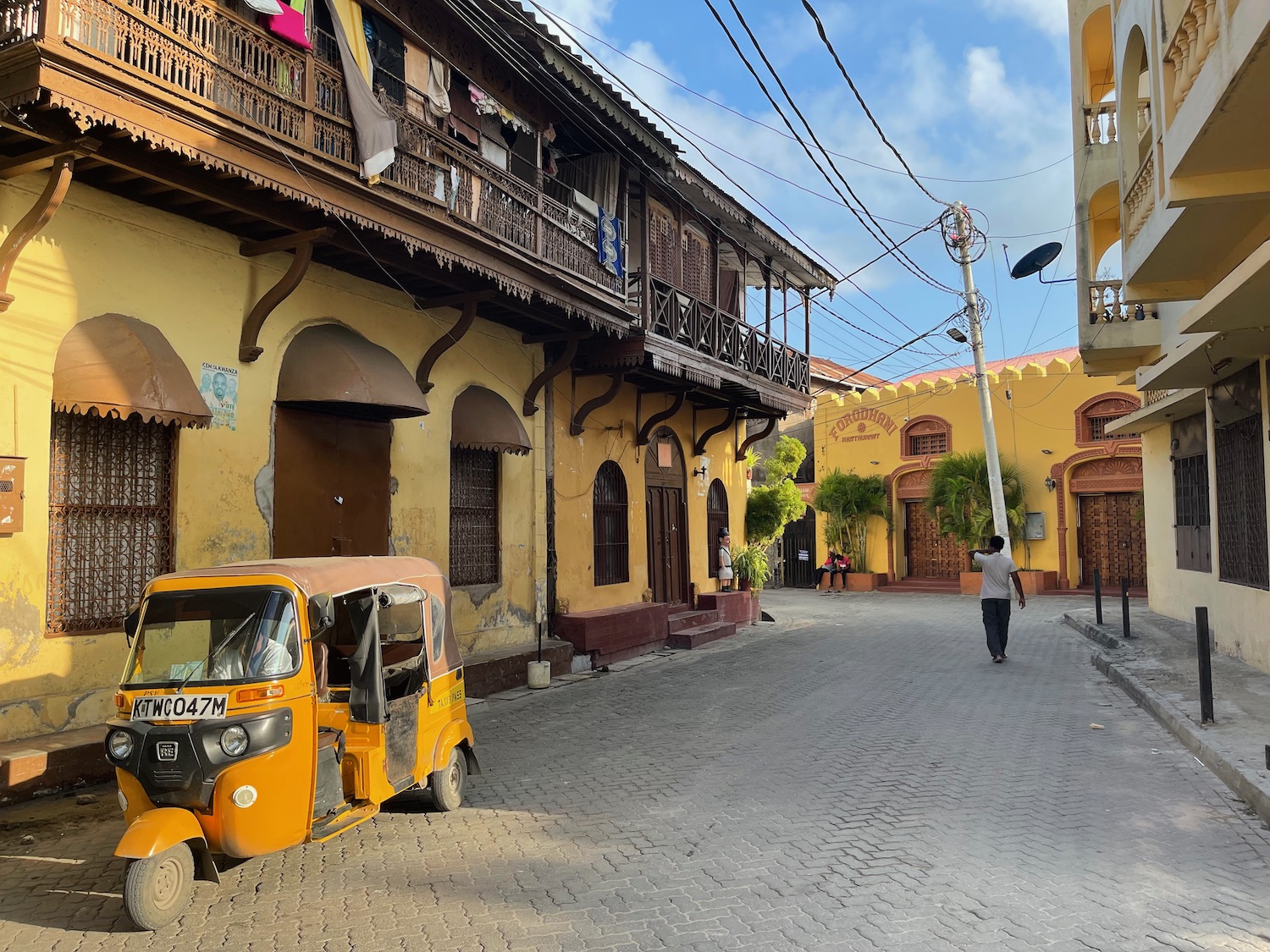 A thin road in the midst of old town Mombasa, Nyali. Features a yellow tuk tuk and a man walking the street.