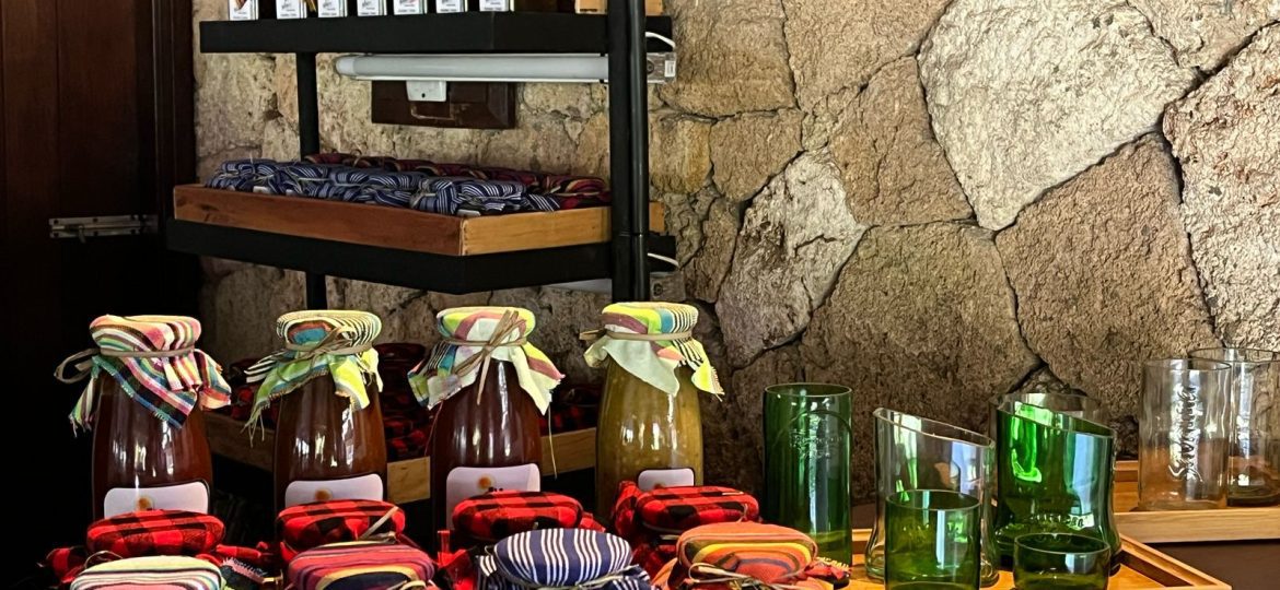 A collection of various empty clear, green glass liquor various sauces and jams, and beer bottles, including Jack Daniel's, Safari, Captain Morgan, and Heineken, neatly arranged on a wooden table, with some bottles repurposed into drinking glasses, showcasing Bahari Beach hotel's sustainability efforts.